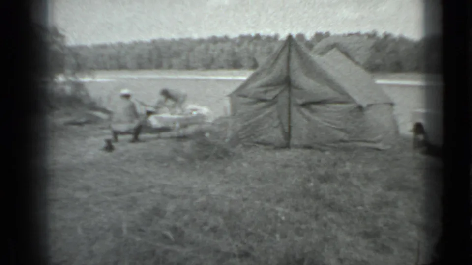 1983 Moscow home-movie frame showing a lakeside tent, seated campers, grass, water, and a wooded far shore.