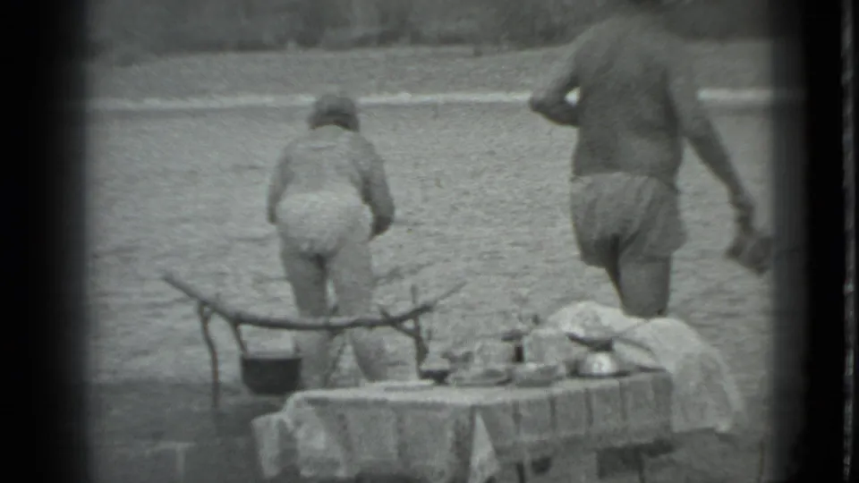 1983 Moscow home-movie frame showing campers at the lakeside, with a table in the foreground and water behind them.