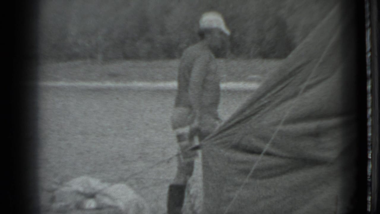 1983 Moscow home-movie frame showing a person beside a canvas tent or tarp in a grassy campground setting.