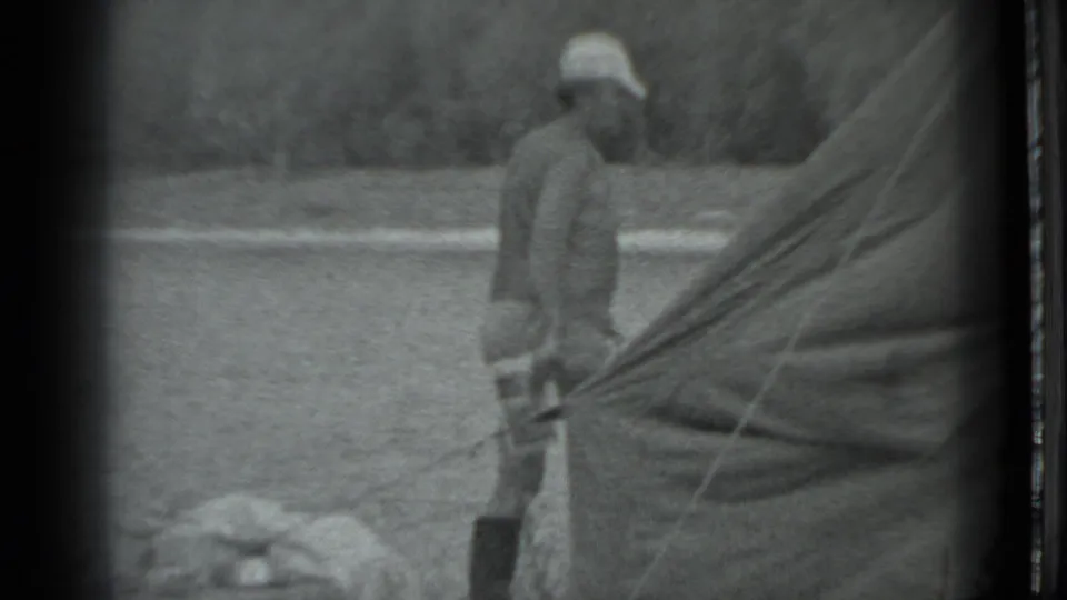 1983 Moscow home-movie frame showing a person beside a canvas tent or tarp in a grassy campground setting.