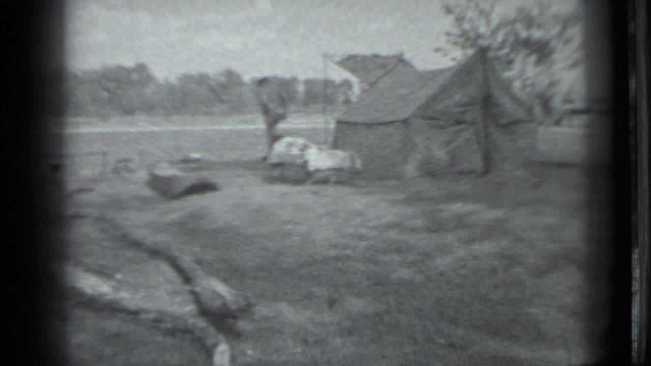 Small riverside campsite with a tent, gear, and grassy bank near a wooded shore.
