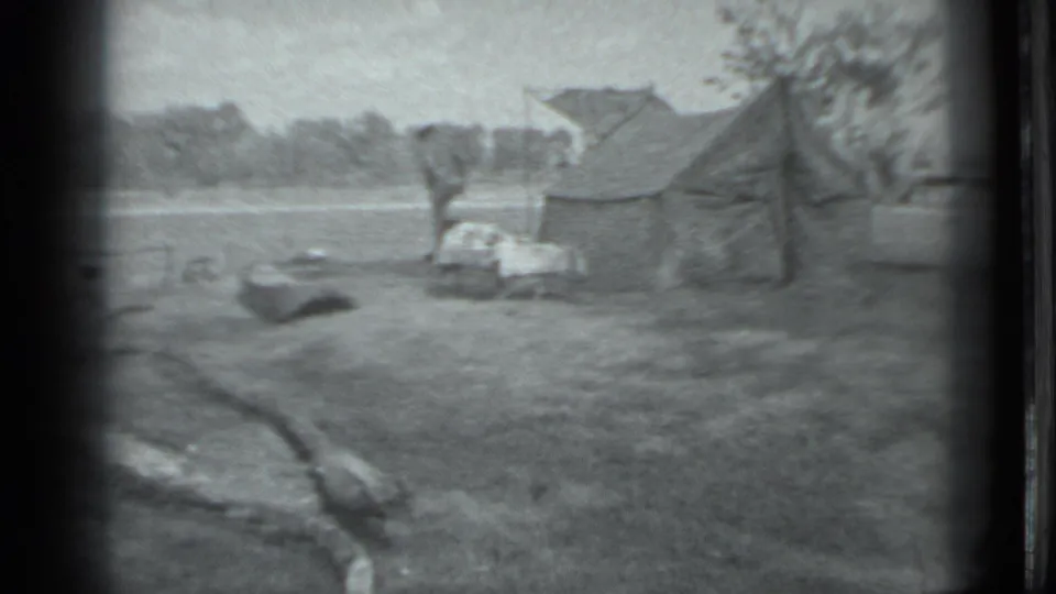 1983 Moscow home-movie frame showing a small riverside camp with a canvas tent, scattered gear, and trees across the water.