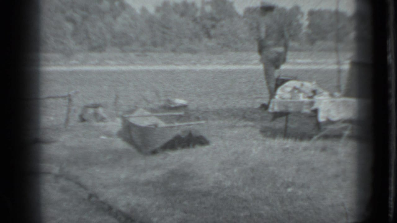 Riverside camp area with low gear, cooking pot, and a figure near a small table.
