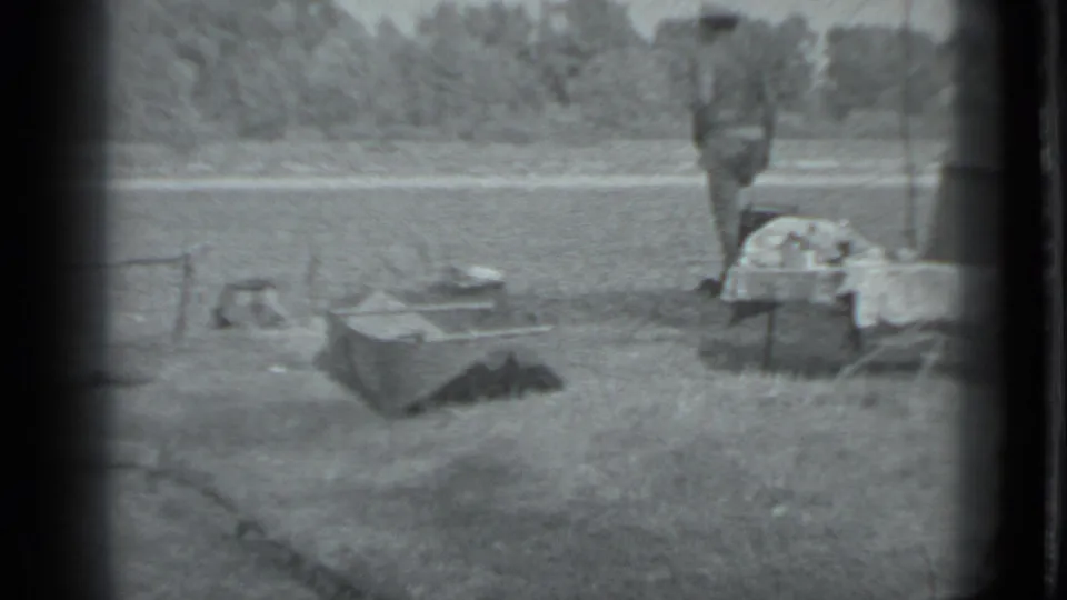 1983 Moscow home-movie frame with a grassy riverbank camp, low cooking gear, and an adult figure near a small table or cot.