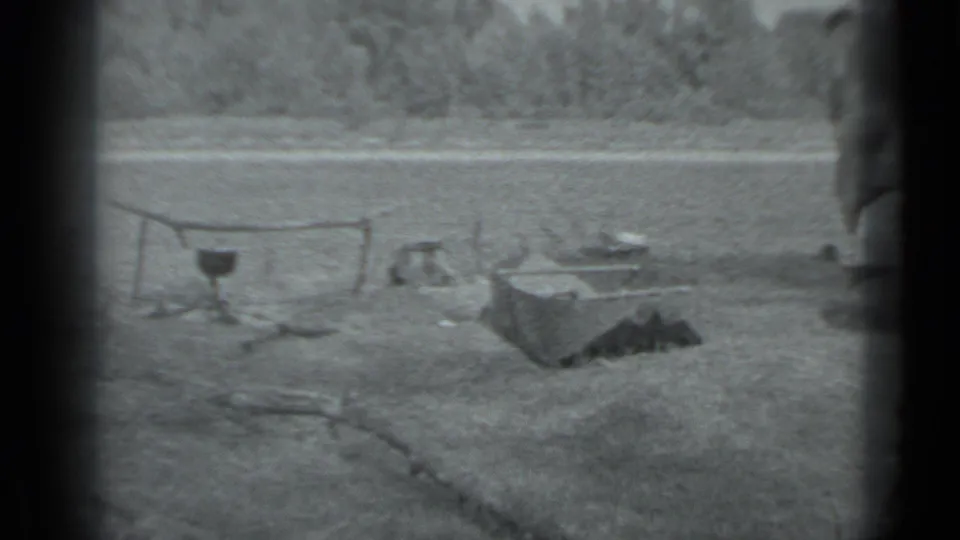 A 1983 Moscow riverside campsite appears with a cooking pot, low camp gear, open grass, and a wooded opposite bank.