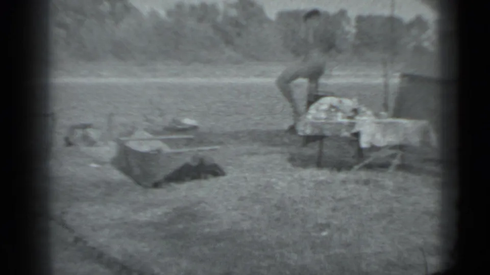 1983 Moscow home-movie frame showing an adult man beside a small riverside camp with a low table or cot and cooking supplies.