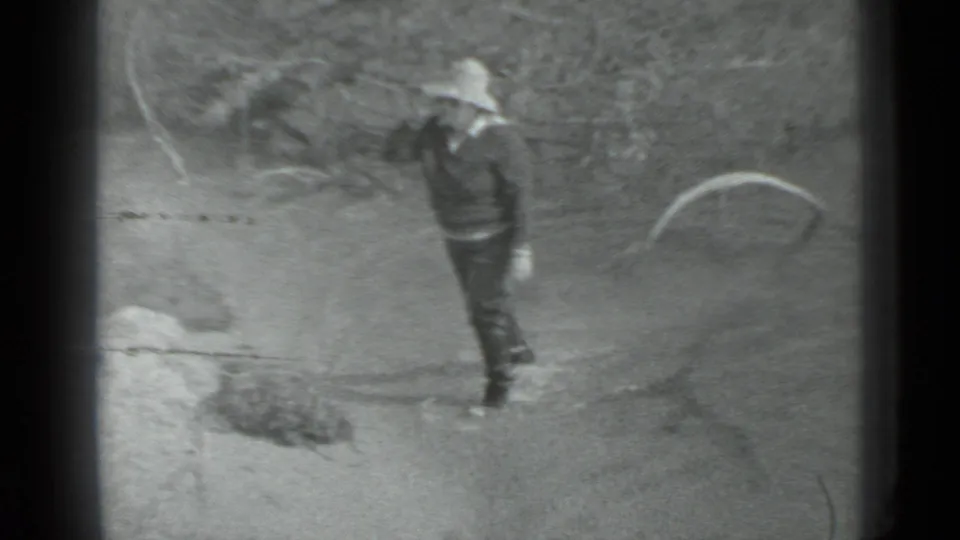 A person in a brimmed hat and dark clothing walks through the wooded camp area in the supplied 1983 Moscow home-movie footage.