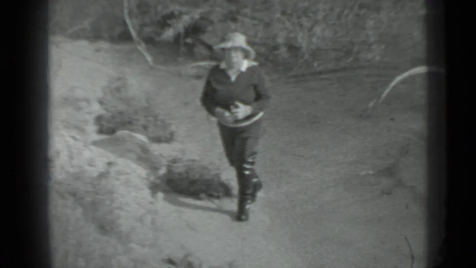 The walking figure remains visible near brush and a curved ground object in the archival 8mm outdoor camp scene.