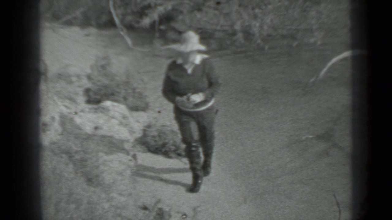 In Moscow, 1983 home-movie footage, a woman in a brimmed hat and dark clothing walks along a sandy shoreline near rocks.
