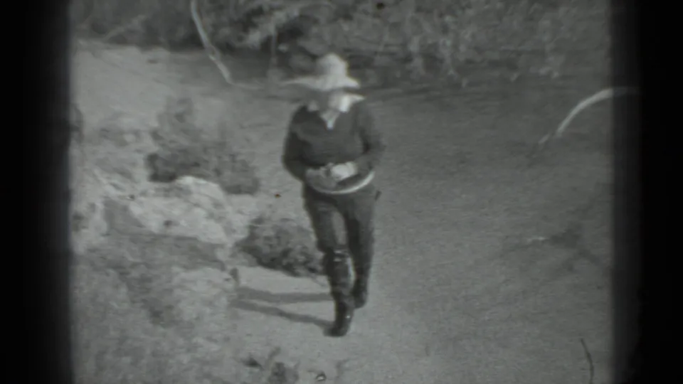 In Moscow, 1983 home-movie footage, a woman in a brimmed hat and dark clothing walks along a sandy shoreline near rocks.
