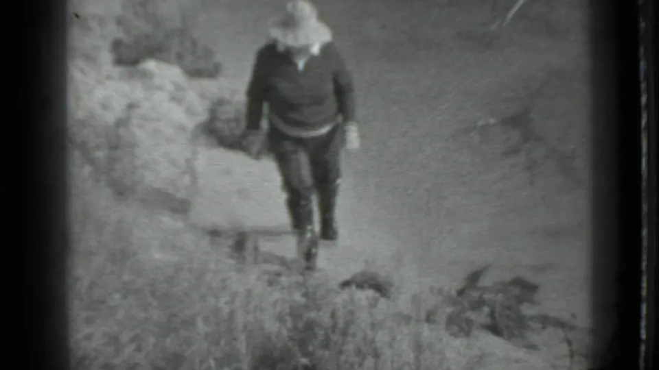 A woman in dark clothing and a pale brimmed hat walks past rocks on a sandy shoreline in the Moscow 1983 clip.