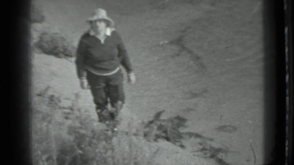 The woman moves closer in the frame, wearing a hat, dark top and boots beside scrub and sandy shoreline ground.