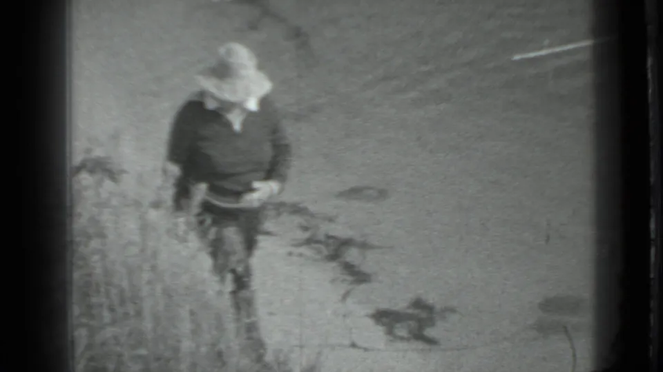 In 1983 Moscow home-movie footage, a person in a wide-brimmed hat walks beside sand and sparse lakeside plants.