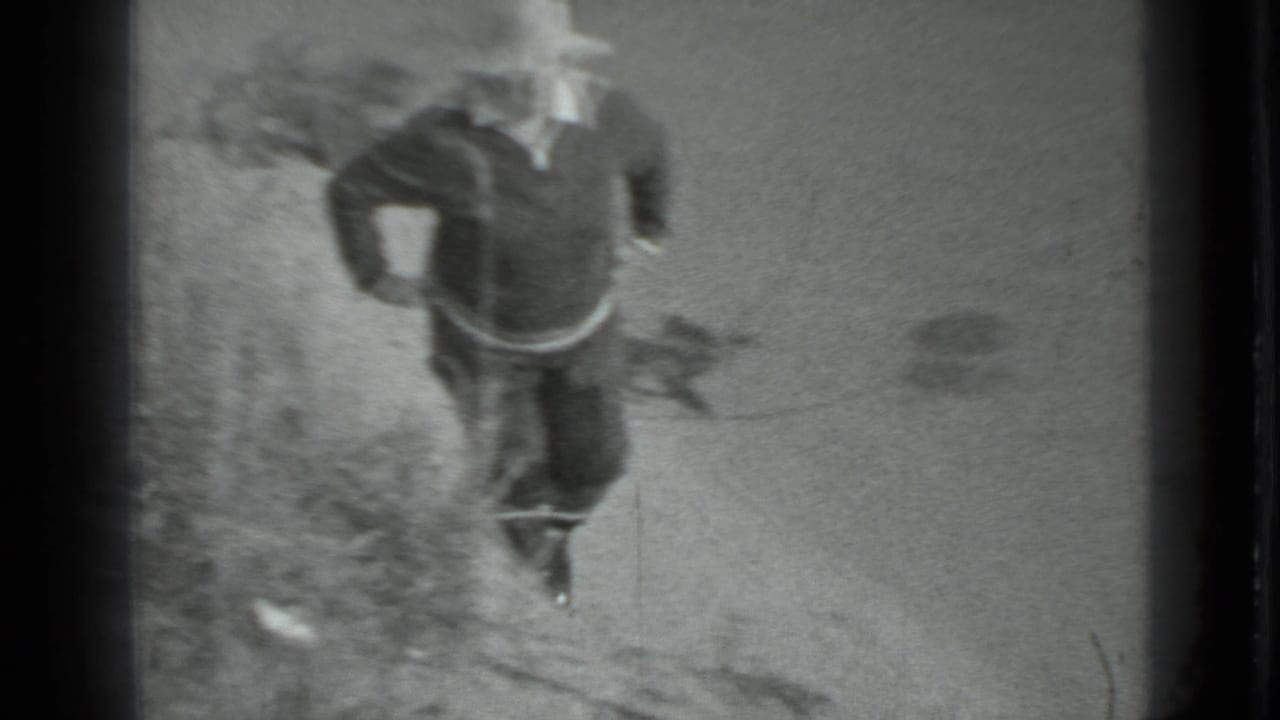 A person in dark clothing and a light wide-brimmed hat moves along a sandy lakeshore in the Moscow 1983 8mm footage.