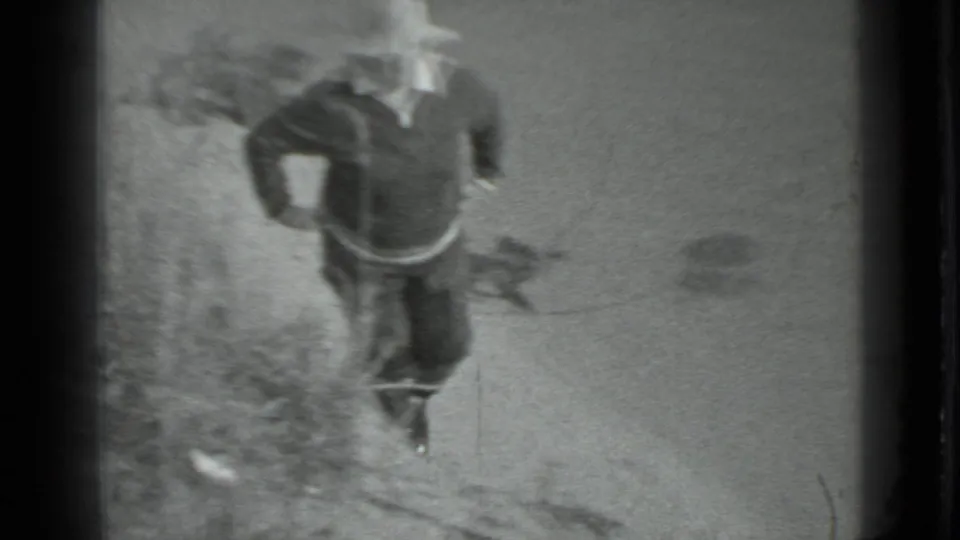 A person in dark clothing and a light wide-brimmed hat moves along a sandy lakeshore in the Moscow 1983 8mm footage.