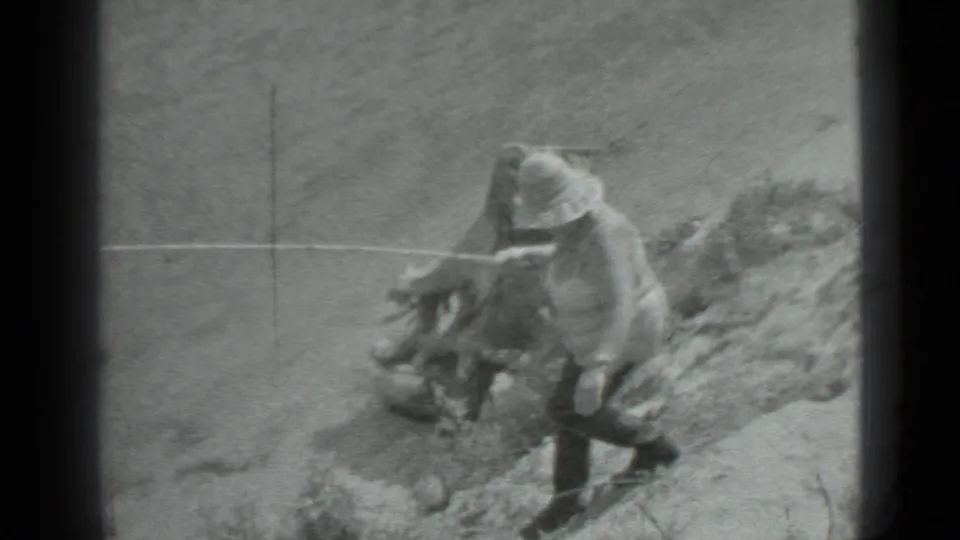 The man leans toward fishing gear or a line on the rocky riverside, framed in grainy black-and-white 8mm footage.