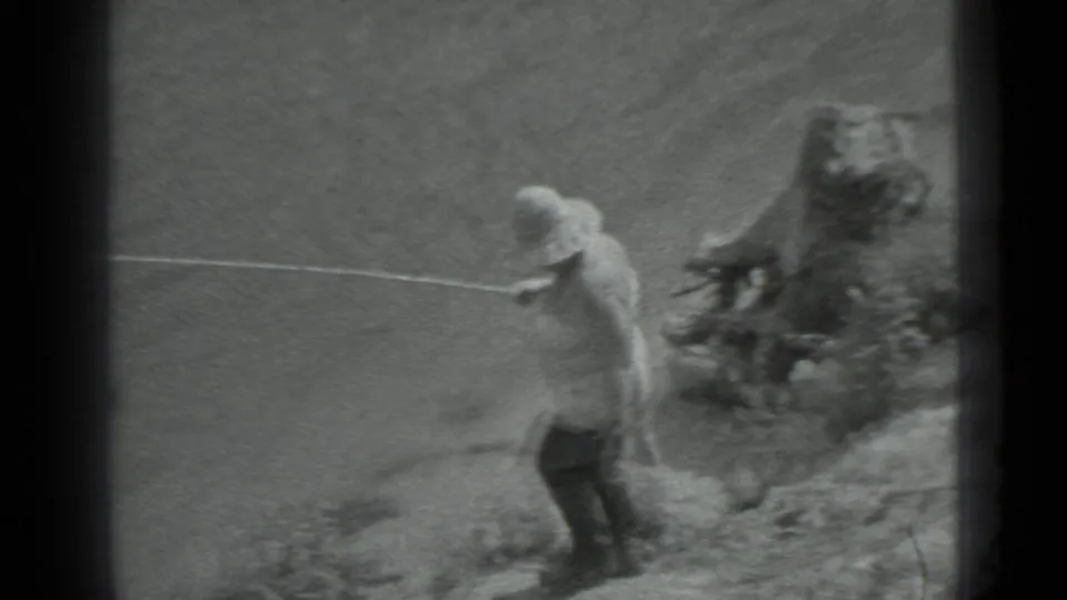 The man holds a long fishing rod or taut line along the riverbank, with water and shoreline rocks visible in the frame.