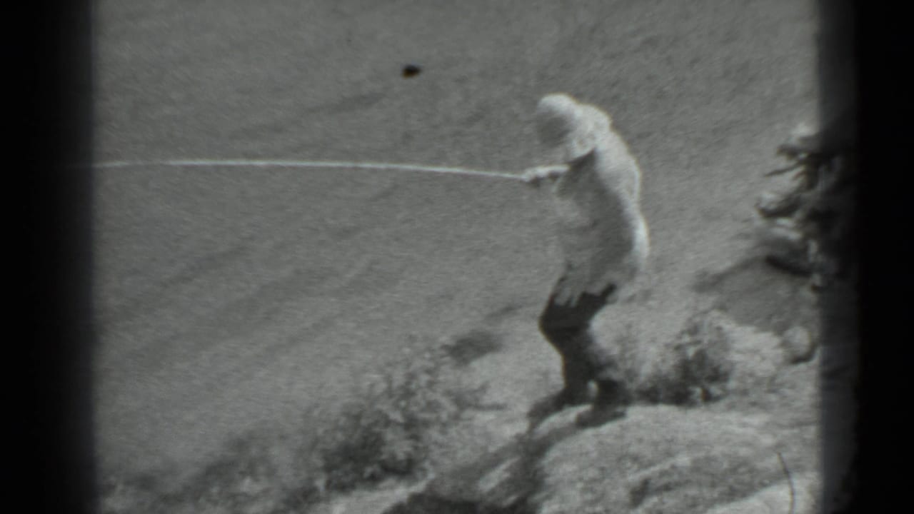 In black-and-white 8mm footage, a woman in a light top and dark pants descends a rocky shoreline with a fishing pole.