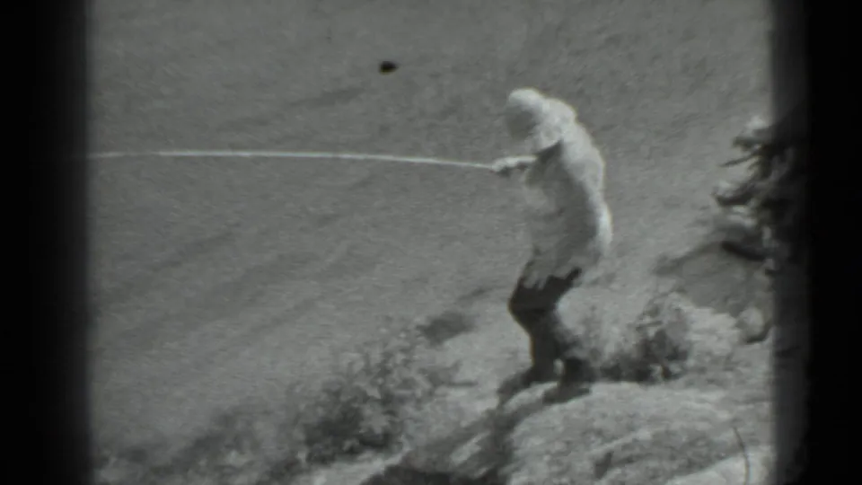 In black-and-white 8mm footage, a woman in a light top and dark pants descends a rocky shoreline with a fishing pole.
