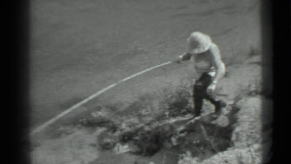 The woman moves along the sloped shore, holding the fishing pole outward as she approaches the waterline.