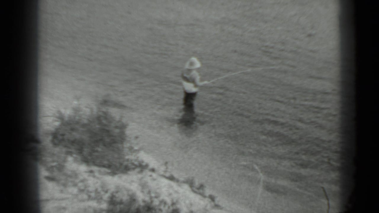 In 1983 Moscow home-movie footage, a man in waders stands in a river with a fishing rod near a low bank.
