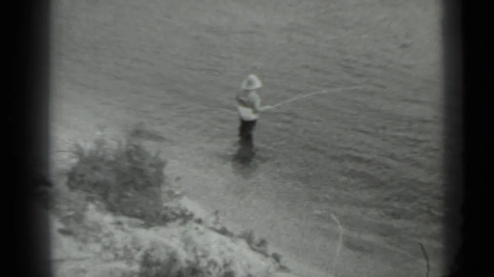 In 1983 Moscow home-movie footage, a man in waders stands in a river with a fishing rod near a low bank.