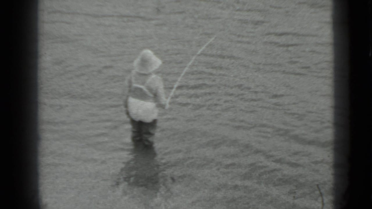 A man in waders is viewed from behind while holding a rod in shallow water in the 1983 Moscow clip.