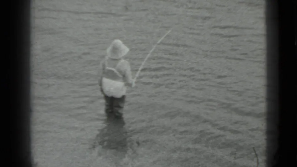 A man in waders is viewed from behind while holding a rod in shallow water in the 1983 Moscow clip.