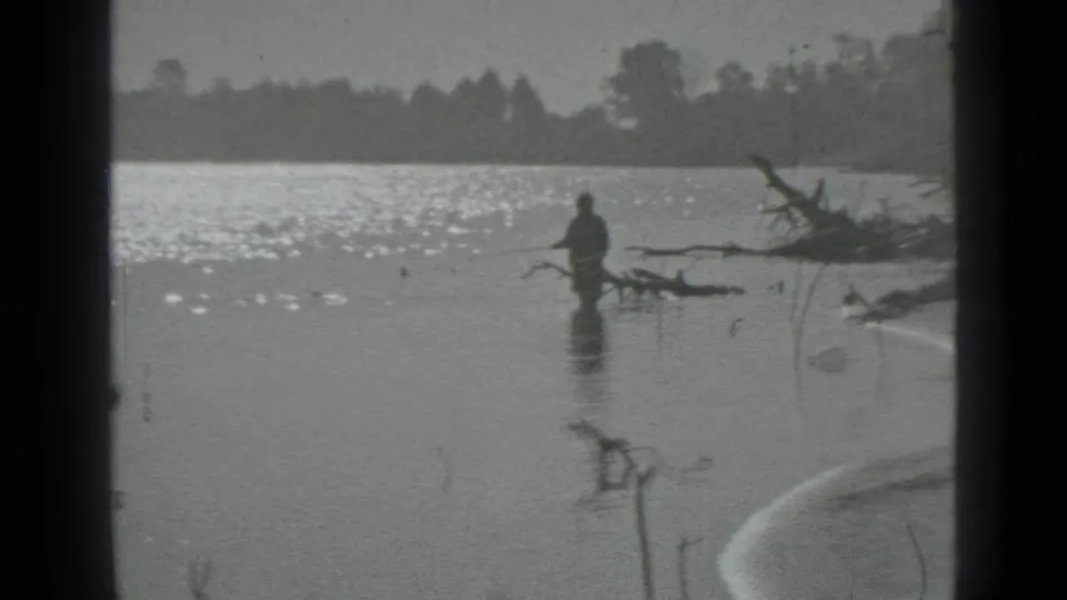 1983 Moscow-area 8mm frame with the fisher seen at a distance in shallow water near branches and sunlit ripples.