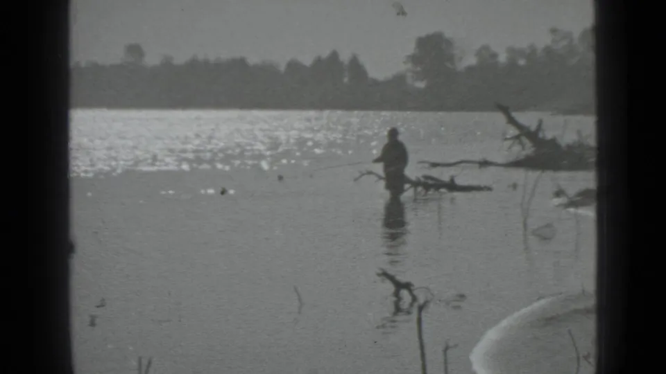 1983 Moscow home-movie frame of a distant man fishing in a calm lake near driftwood and a tree-lined shore.