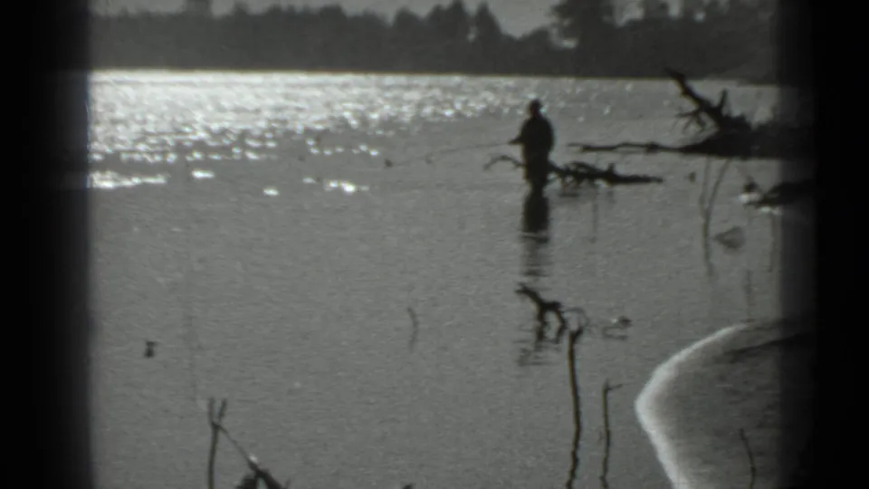 1983 Moscow home-movie frame showing a distant fisherman in reflective lake water near driftwood and shoreline reeds.