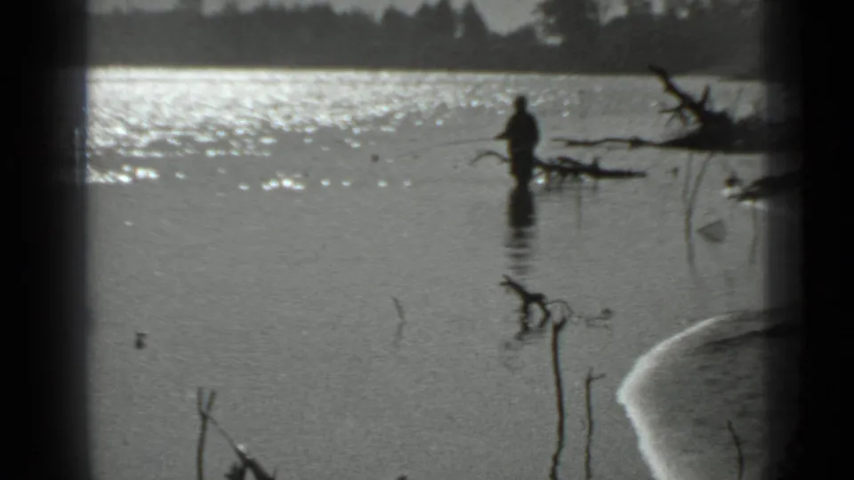 A distant man appears to fish while standing waist-deep in a calm Moscow-area lake in 1983 home-movie footage.