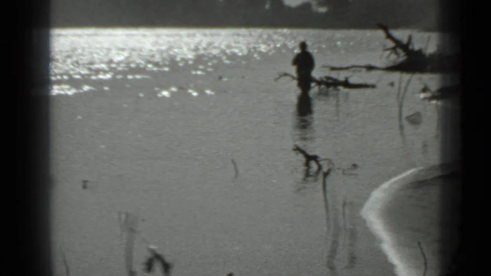 The fisherman remains in the lake, partly silhouetted against bright water, with reeds and driftwood along the frame edge.