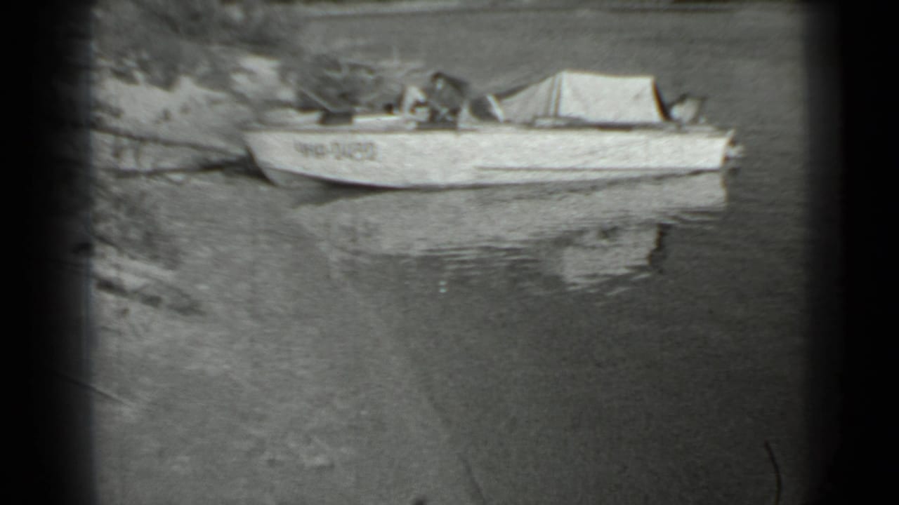 The camera holds on the moored covered boat by the lakeshore, with rippled reflections and soft 8mm framing.