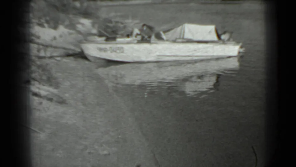 The camera holds on the moored covered boat by the lakeshore, with rippled reflections and soft 8mm framing.