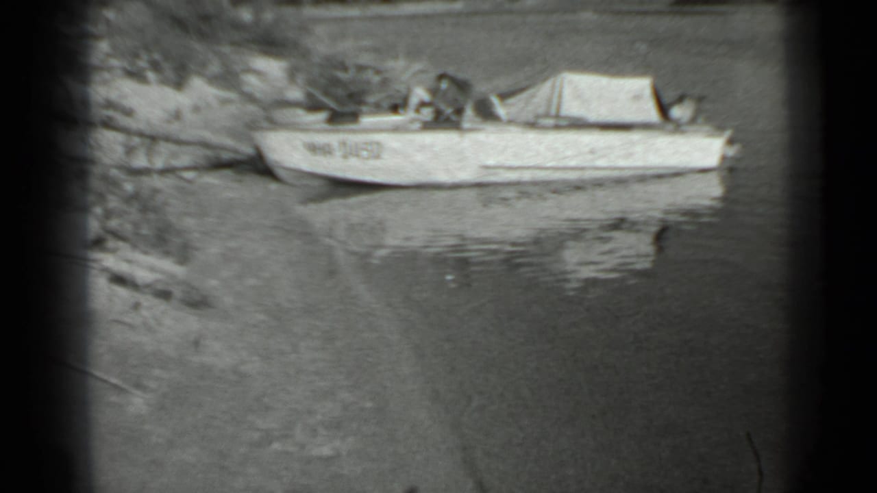 Small aluminum boat near a Moscow-area shoreline in 1983 home-movie footage, with calm water and sparse vegetation visible.