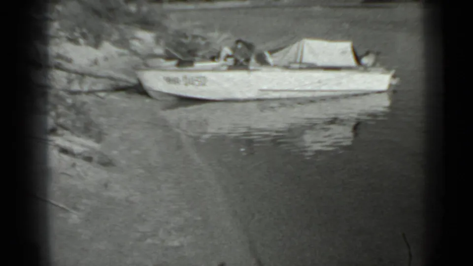 Small aluminum boat near a Moscow-area shoreline in 1983 home-movie footage, with calm water and sparse vegetation visible.