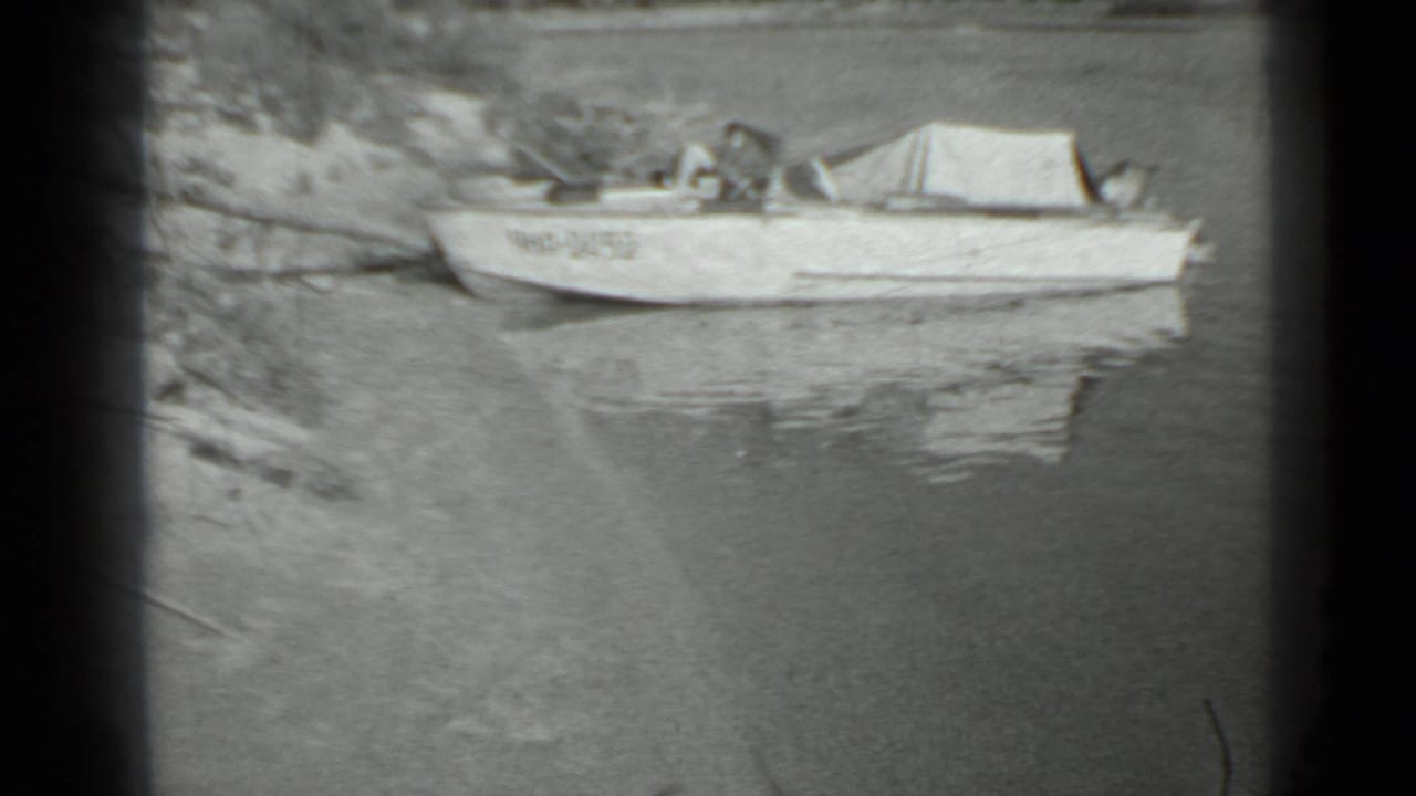 Aluminum boat anchored near shore in 1983 Moscow home-movie footage, with faint markings and reflections on still water.