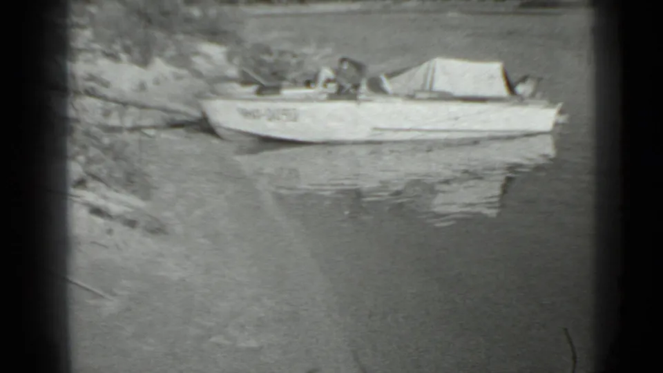 Aluminum boat anchored near shore in 1983 Moscow home-movie footage, with faint markings and reflections on still water.