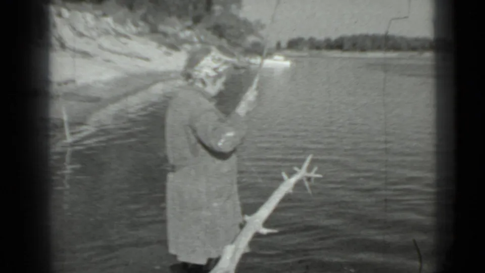 Older woman in a long coat and head covering fishes from a Moscow-area shore in 1983 home-movie footage.
