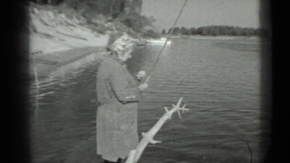 Older woman fishes from a shoreline in Moscow-area 1983 footage, with open water, a branch, and distant trees behind her.