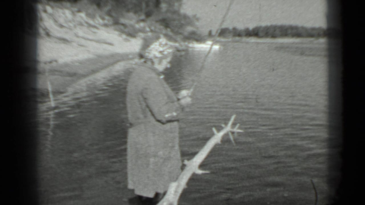 Woman in a long coat and headscarf fishing beside rippled lake water.