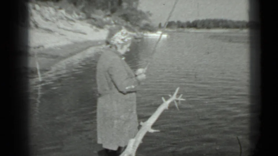 In 1983 Moscow home-movie footage, a woman in a long coat and patterned headscarf stands at a lake edge holding a fishing rod.