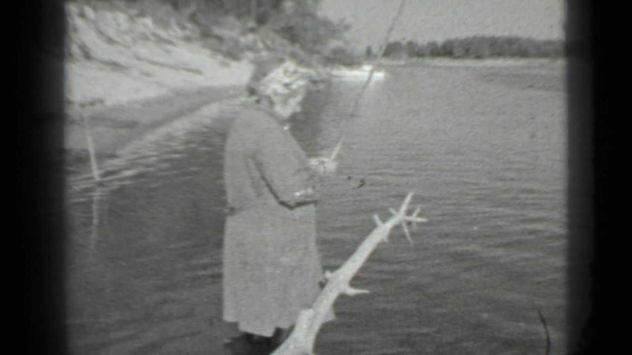 A woman stands side-on by a Moscow lake in 1983, holding a fishing rod as wind-textured water fills the background.