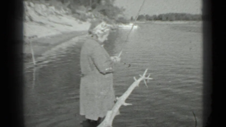 A woman stands side-on by a Moscow lake in 1983, holding a fishing rod as wind-textured water fills the background.