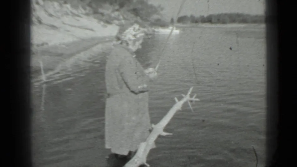 Black-and-white 8mm frame of a woman fishing from a lakeshore near Moscow, with rippled water, sand, and distant trees visible.