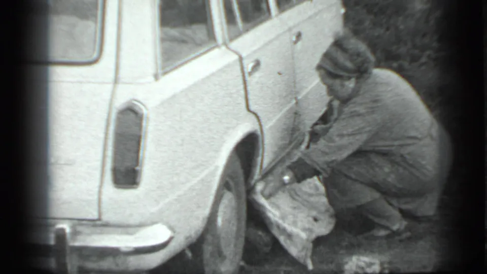 In the Moscow 1983 8mm footage, a woman in a coat and headscarf crouches by a light-colored station wagon, appearing to work near the rear wheel.