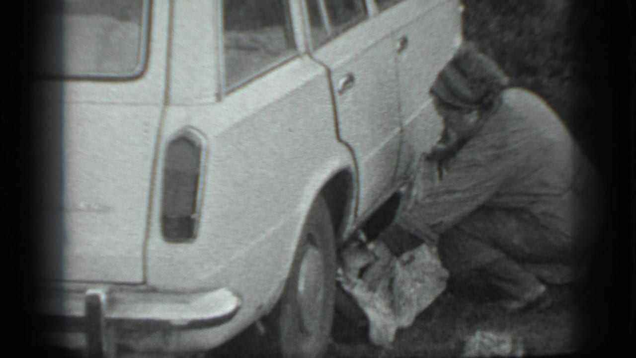 In Moscow 1983 footage, a man in a dark coat crouches by a white station wagon's rear wheel with a cloth or bag on the ground.