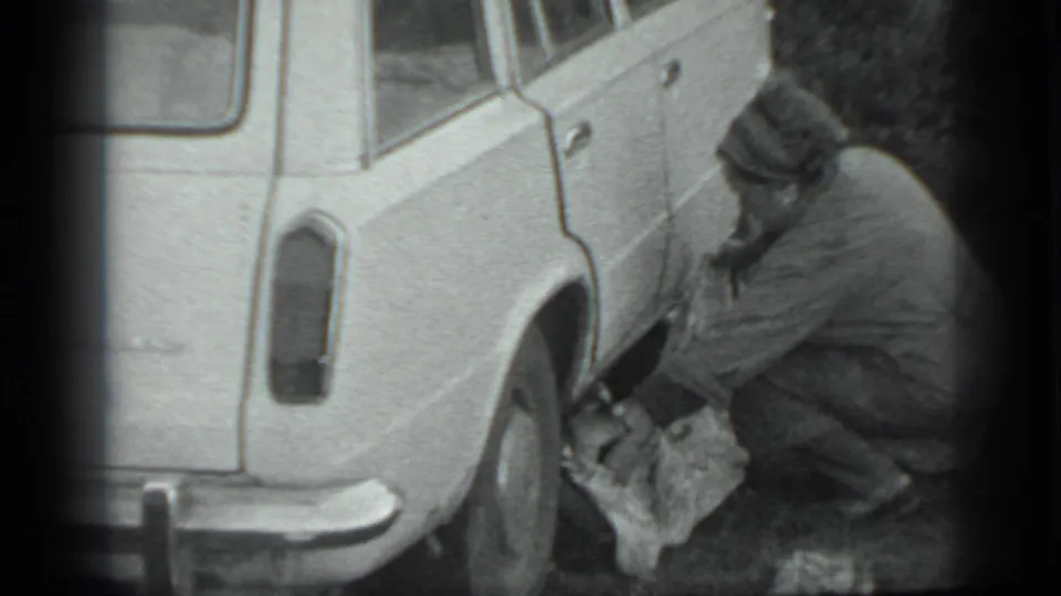In Moscow 1983 footage, a man in a dark coat crouches by a white station wagon's rear wheel with a cloth or bag on the ground.