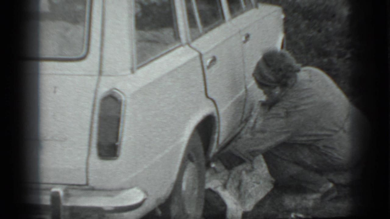 The man remains crouched beside the station wagon in Moscow, handling a cloth-covered item near the rear wheel.